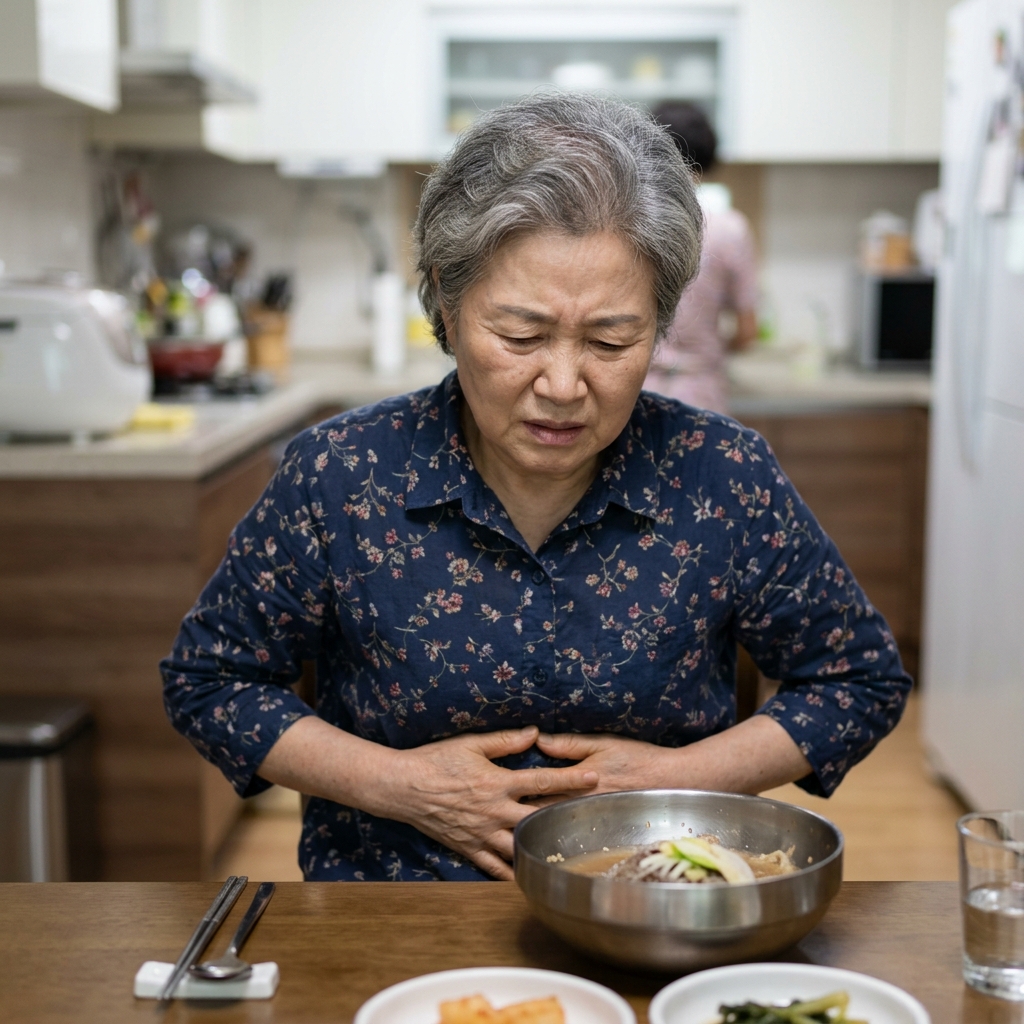 70대 여성이 식탁에서 냉면 그릇 앞에 앉아 배를 손으로 감싸 쥐고 있는 모습, 실내 조명 아래 불편한 표정